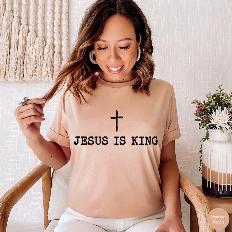 K&ouml;nnte beinhalten: Ein pfirsichfarbenes T-Shirt mit dem Text "JESUS IS KING" in Schwarz aufgedruckt, mit einem einfachen Kreuz &uuml;ber dem Text. Das Shirt hat kurze &Auml;rmel und einen Rundhalsausschnitt. Das Model sitzt auf einem Holzstuhl.