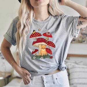 K&ouml;nnte beinhalten: Hellgraues T-Shirt mit einer skurrilen Illustration. Das Design zeigt rote Pilze mit wei&szlig;en Punkten, einen gelben Schmetterling, eine rot getigerte Katze und G&auml;nsebl&uuml;mchen. Ein l&auml;ssiges Kleidungsst&uuml;ck f&uuml;r den Alltag.