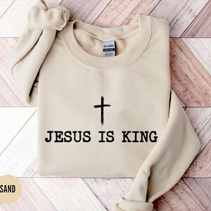 K&ouml;nnte beinhalten: Sandfarbener Pullover mit dem schwarzen Text "JESUS IS KING" und einem schwarzen Kreuz. Der Pullover ist gefaltet, wobei die &Auml;rmel und der Kragen sichtbar sind. Der Hintergrund hat ein helles Holzmuster.