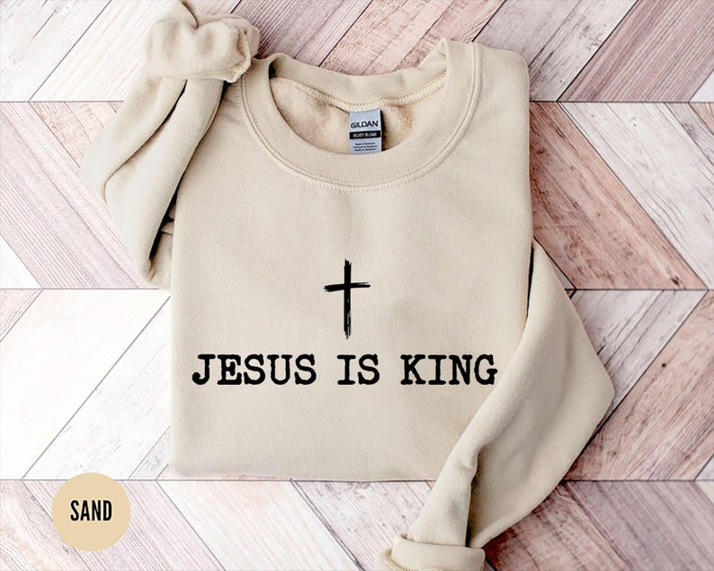 K&ouml;nnte beinhalten: Sandfarbener Pullover mit dem schwarzen Text "JESUS IS KING" und einem schwarzen Kreuz. Der Pullover ist gefaltet, wobei die &Auml;rmel und der Kragen sichtbar sind. Der Hintergrund hat ein helles Holzmuster.