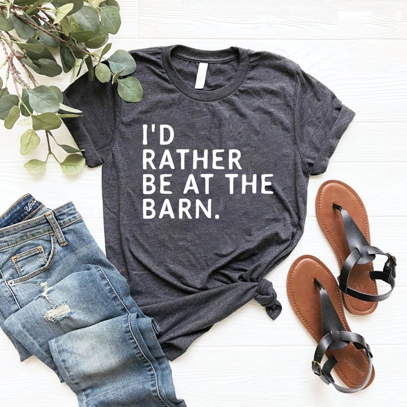 Puede incluir: Camiseta gris oscuro con el texto "I'D RATHER BE AT THE BARN." La camiseta se combina con vaqueros azules y sandalias marrones. Ramas de hojas verdes est&aacute;n dispuestas en la parte superior izquierda de la imagen.