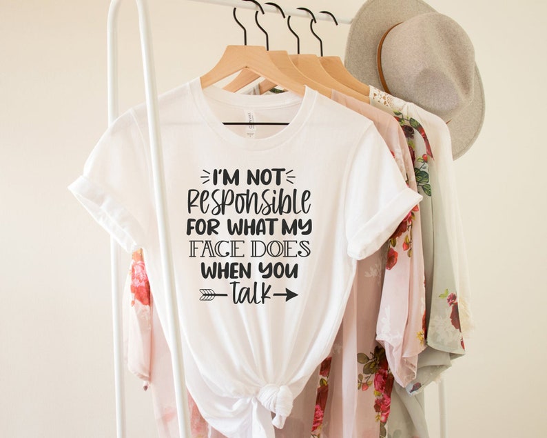 以下が含まれることがあります： I'M NOT RESPONSIBLE FOR WHAT MY FACE DOES WHEN YOU TALKという黒いテキストがプリントされた白い半袖Tシャツ。グラフィックデザインの上下の行には、装飾的な矢印のシンボルがあしらわれています。