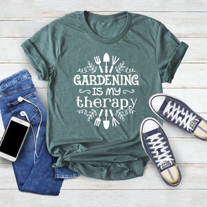 Op de afbeelding: Een tealgroen T-shirt met de tekst "Gardening is my therapy" in wit. De tekst wordt omringd door een bloemmotief met tuingereedschap.