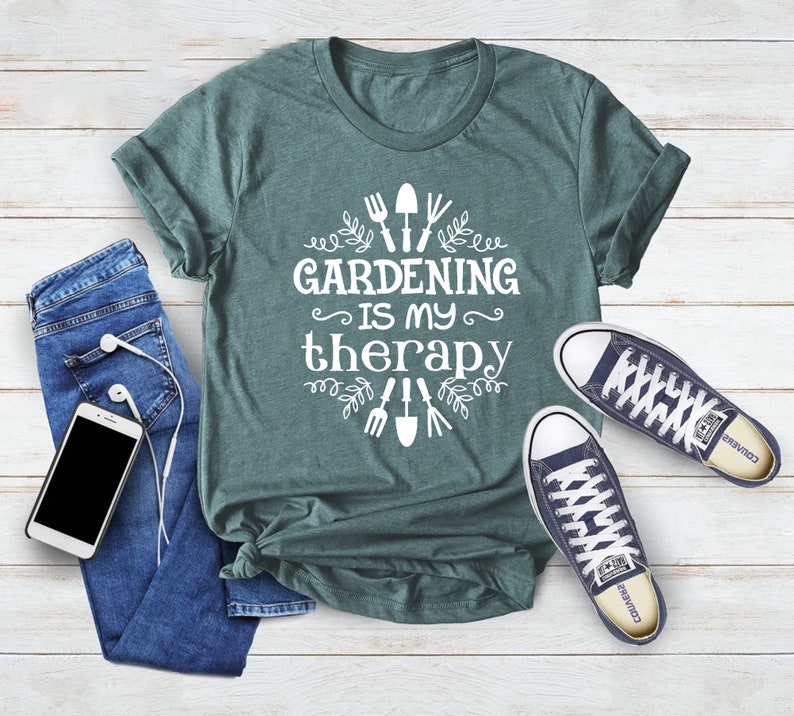 Op de afbeelding: Een tealgroen T-shirt met de tekst "Gardening is my therapy" in wit. De tekst wordt omringd door een bloemmotief met tuingereedschap.