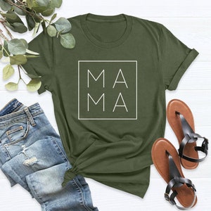 May include: Olive green t-shirt with the word "MAMA" in a white square. The shirt is paired with blue jeans and black sandals. The t-shirt is a casual, everyday wear item.