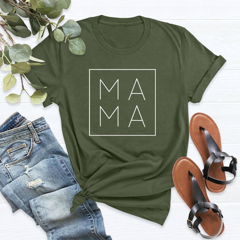 May include: Olive green t-shirt with the word "MAMA" in a white square. The shirt is paired with blue jeans and black sandals. The t-shirt is a casual, everyday wear item.