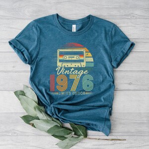 May include: A heathered blue short-sleeve t-shirt featuring a graphic of a retro cassette tape above the text Vintage 1976 LIMITED EDITION. The design incorporates a sunset motif with horizontal stripes in red, orange, yellow, and teal colours.
