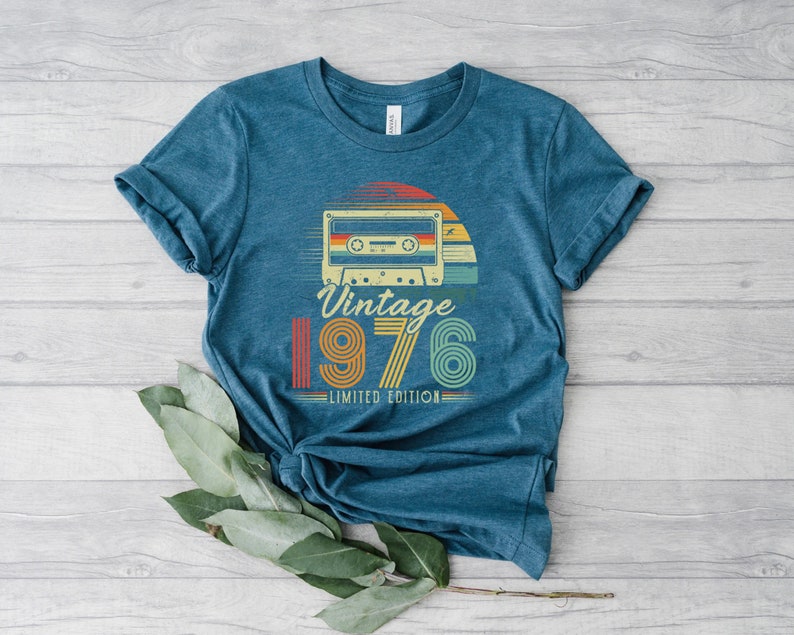 May include: A heathered blue short-sleeve t-shirt featuring a graphic of a retro cassette tape above the text Vintage 1976 LIMITED EDITION. The design incorporates a sunset motif with horizontal stripes in red, orange, yellow, and teal colours.