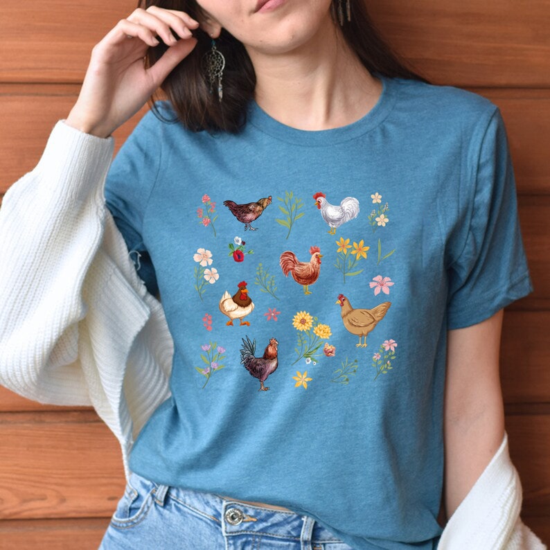 May include: A teal blue t-shirt with a colorful graphic print of chickens and flowers. The graphic features a variety of chickens in different poses, including a rooster, a hen, and chicks. The flowers are in shades of pink, yellow, and purple.