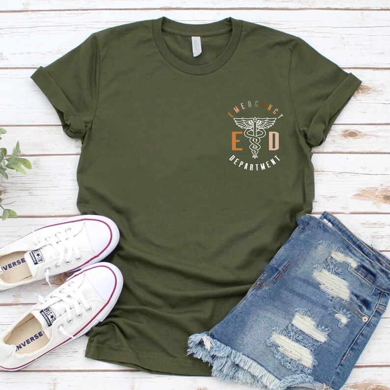 May include: Olive green t-shirt with rolled sleeves, featuring a small graphic on the chest that reads "Emergency Department" with a medical symbol. The shirt is paired with denim shorts and white canvas sneakers.
