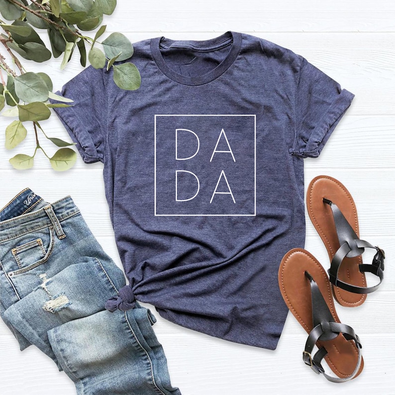 May include: A navy blue t-shirt with a white square graphic that says "DA DA" on it. The shirt is laying on a white surface with a pair of brown sandals and blue jeans.