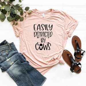 May include: A light pink t-shirt with the text "EASILY DISTRACTED BY COWS" in black. The shirt is paired with blue jeans and brown sandals. A sprig of greenery is in the background.