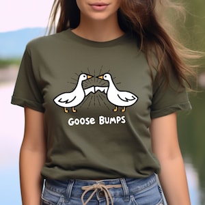 Puede incluir: Una camiseta verde con una ilustraci&oacute;n de dibujos animados de dos gansos blancos que se miran con el texto "Goose Bumps" debajo.