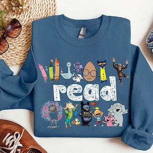 May include: An indigo blue crewneck sweatshirt featuring a graphic print of various colorful cartoon characters and the word "read" in white, textured lettering. The design includes illustrations of crayons, a pigeon, a cat, a monkey, and an elephant.