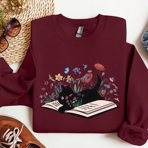 May include: A maroon sweatshirt with a black cat reading a book surrounded by flowers and butterflies. The book is open and the cat is looking at the book.