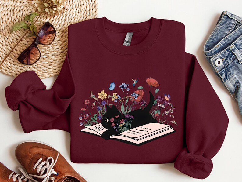 May include: A maroon sweatshirt with a black cat reading a book surrounded by flowers and butterflies. The book is open and the cat is looking at the book.