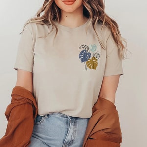 May include: Beige t-shirt with a small, colorful botanical illustration on the chest. The design features stylized leaves in shades of blue, green, and gold. The model is wearing blue jeans and a brown jacket.
