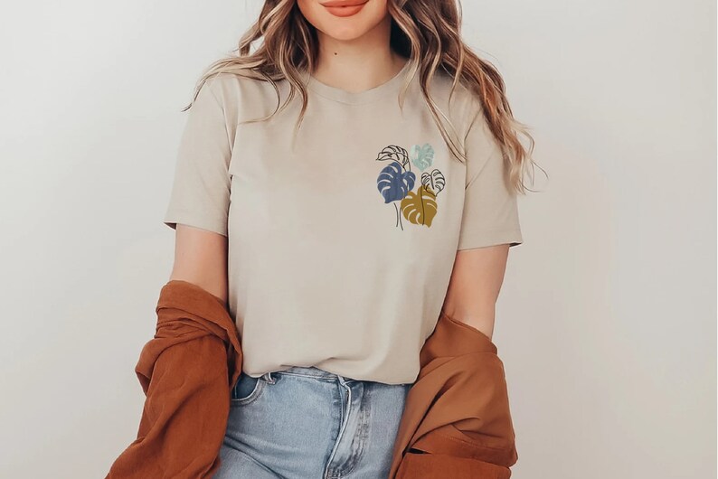 May include: Beige t-shirt with a small, colorful botanical illustration on the chest. The design features stylized leaves in shades of blue, green, and gold. The model is wearing blue jeans and a brown jacket.