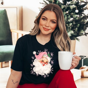 May include: Black t-shirt featuring a vintage Santa Claus graphic with a pink hat and white beard, surrounded by snowflakes. The Santa Claus graphic is in the center of the shirt. The person is wearing red pants and holding a white mug.