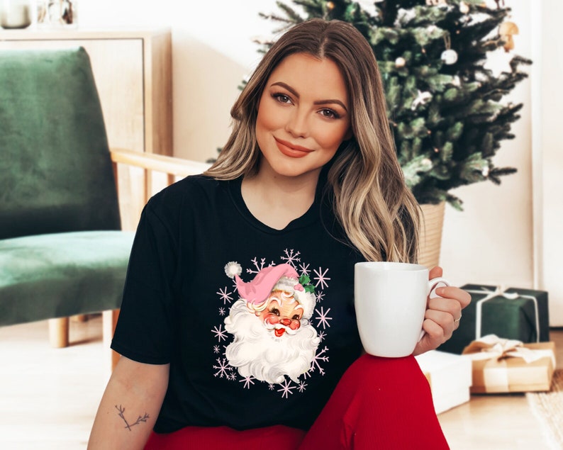 May include: Black t-shirt featuring a vintage Santa Claus graphic with a pink hat and white beard, surrounded by snowflakes. The Santa Claus graphic is in the center of the shirt. The person is wearing red pants and holding a white mug.