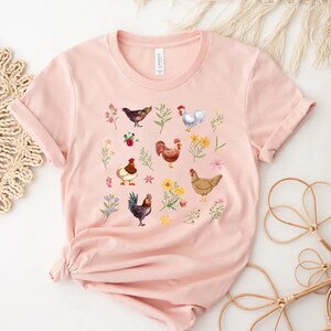 May include: A pink t-shirt with a graphic print of chickens and flowers. The chickens are in various colors, including brown, white, and black. The flowers are in shades of pink, yellow, and purple.