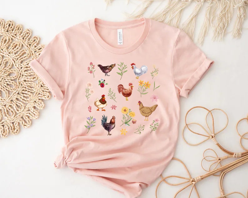 May include: A pink t-shirt with a graphic print of chickens and flowers. The chickens are in various colors, including brown, white, and black. The flowers are in shades of pink, yellow, and purple.