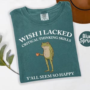 May include: A blue spruce colored t-shirt featuring a graphic of a standing frog holding a small orange mug. The text reads WISH I LACKED CRITICAL THINKING SKILLS above the frog and Y'ALL SEEM SO HAPPY below the frog in white lettering.