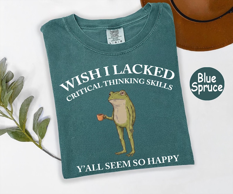 May include: A blue spruce colored t-shirt featuring a graphic of a standing frog holding a small orange mug. The text reads WISH I LACKED CRITICAL THINKING SKILLS above the frog and Y'ALL SEEM SO HAPPY below the frog in white lettering.