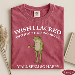 May include: A crimson short-sleeve t-shirt featuring a graphic of a green frog holding a small brown mug. The white text reads WISH I LACKED CRITICAL THINKING SKILLS and Y'ALL SEEM SO HAPPY. The shirt has a crew neckline and a Comfort Colors brand label.