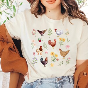 May include: A white t-shirt with a colorful pattern of chickens and flowers. The chickens are brown, white, and red with different patterns. The flowers are pink, yellow, and purple.