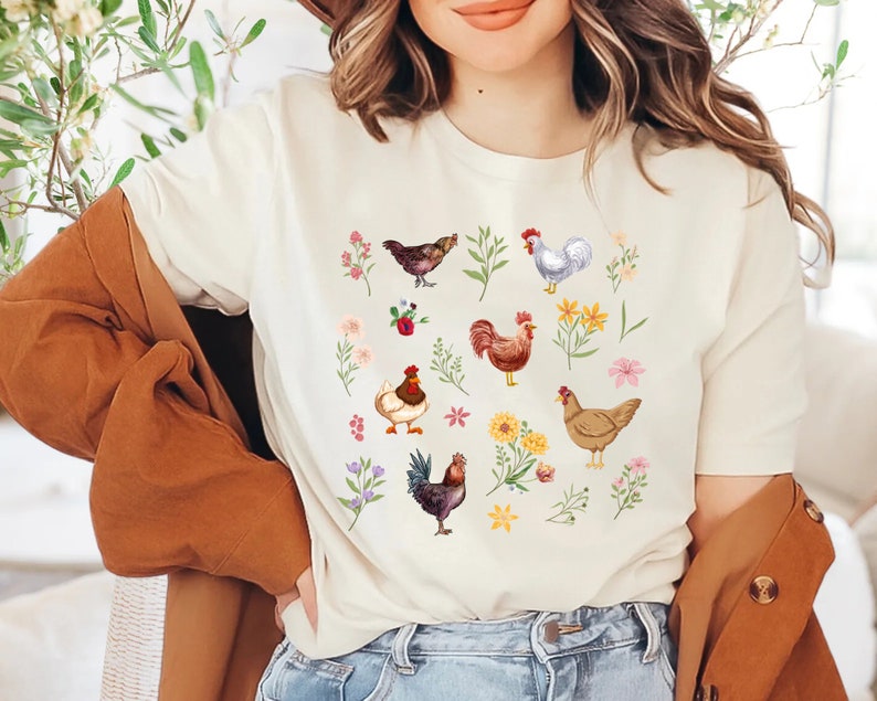 May include: A white t-shirt with a colorful pattern of chickens and flowers. The chickens are brown, white, and red with different patterns. The flowers are pink, yellow, and purple.