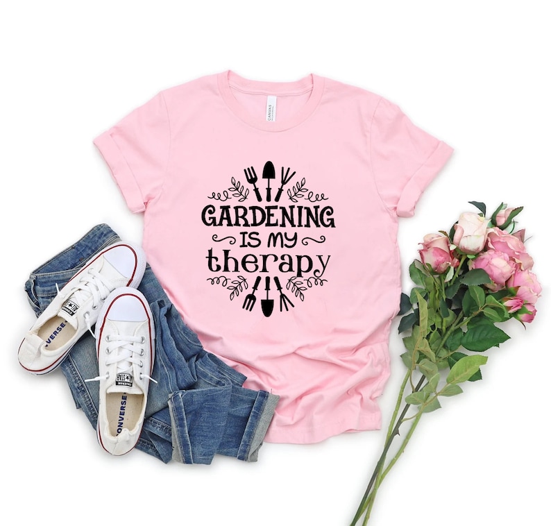 Op de afbeelding: Roze T-shirt met een zwarte afbeelding met de tekst "Tuinieren is mijn therapie" met tuingereedschap en bladeren rond de tekst.