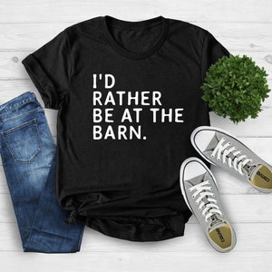 Puede incluir: Camiseta negra con el texto blanco "I'D RATHER BE AT THE BARN." La camiseta se combina con vaqueros azules y zapatillas grises. Una peque&ntilde;a planta verde tambi&eacute;n est&aacute; en la imagen. La camiseta es una prenda informal.