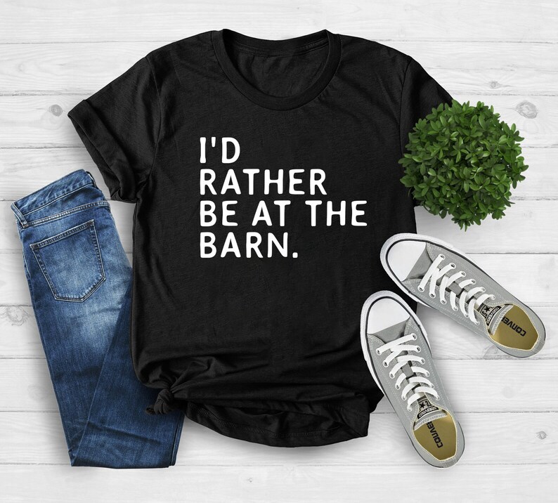 Puede incluir: Camiseta negra con el texto blanco "I'D RATHER BE AT THE BARN." La camiseta se combina con vaqueros azules y zapatillas grises. Una peque&ntilde;a planta verde tambi&eacute;n est&aacute; en la imagen. La camiseta es una prenda informal.