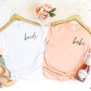 K&ouml;nnte beinhalten: Zwei T-Shirts, eines wei&szlig; und eines pfirsichfarben, werden auf goldenen Kleiderb&uuml;geln pr&auml;sentiert. Das wei&szlig;e Shirt tr&auml;gt den Schriftzug "bride" in schwarzer Schrift, und das pfirsichfarbene Shirt tr&auml;gt den Schriftzug "babe". Zubeh&ouml;r umfasst Schuhe, Blumen und eine Flasche Sekt.