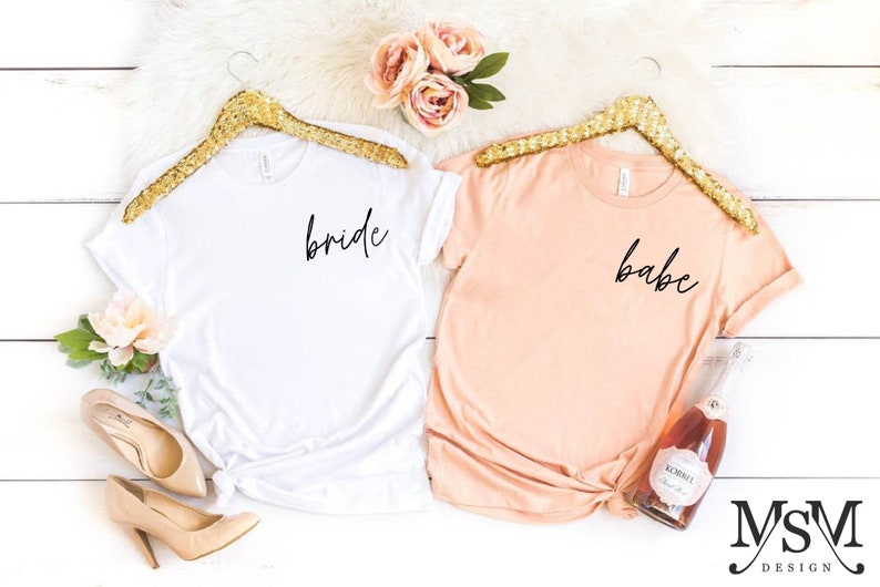 K&ouml;nnte beinhalten: Zwei T-Shirts, eines wei&szlig; und eines pfirsichfarben, werden auf goldenen Kleiderb&uuml;geln pr&auml;sentiert. Das wei&szlig;e Shirt tr&auml;gt den Schriftzug "bride" in schwarzer Schrift, und das pfirsichfarbene Shirt tr&auml;gt den Schriftzug "babe". Zubeh&ouml;r umfasst Schuhe, Blumen und eine Flasche Sekt.