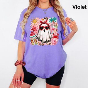 Puede incluir: Una camiseta violeta con un estampado gr&aacute;fico de un fantasma que lleva un pa&ntilde;uelo rojo y gafas de sol en forma de coraz&oacute;n. El dise&ntilde;o incluye elementos veraniegos como una palmera, un c&oacute;ctel, sand&iacute;a, estrella de mar, cangrejo y chanclas. El texto Violet es visible.