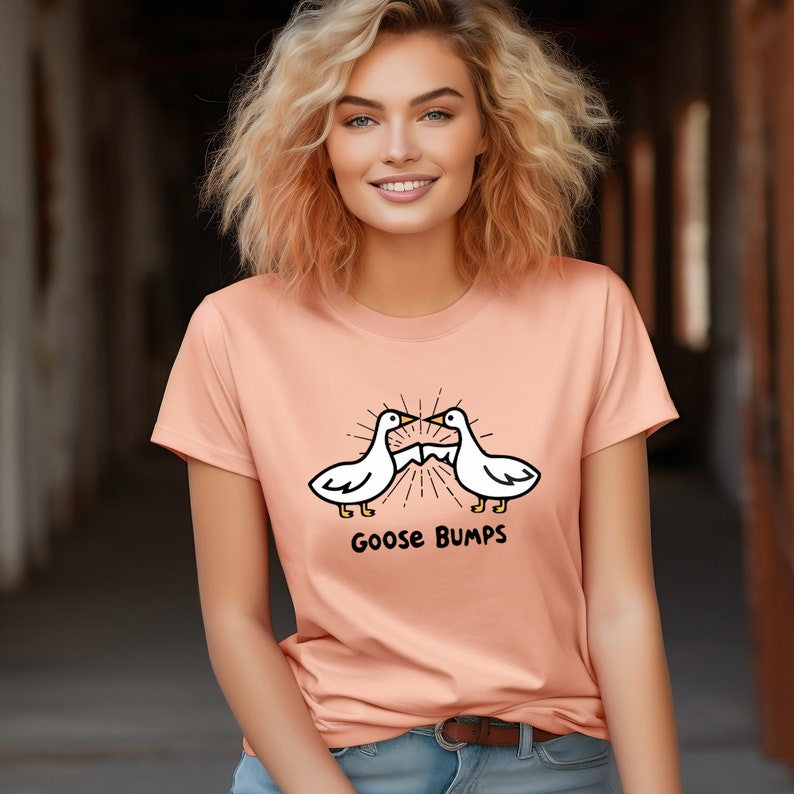 Puede incluir: Una camiseta de color melocot&oacute;n con una imagen de dibujos animados blanca de dos gansos que se miran con el texto "Goose Bumps" debajo.
