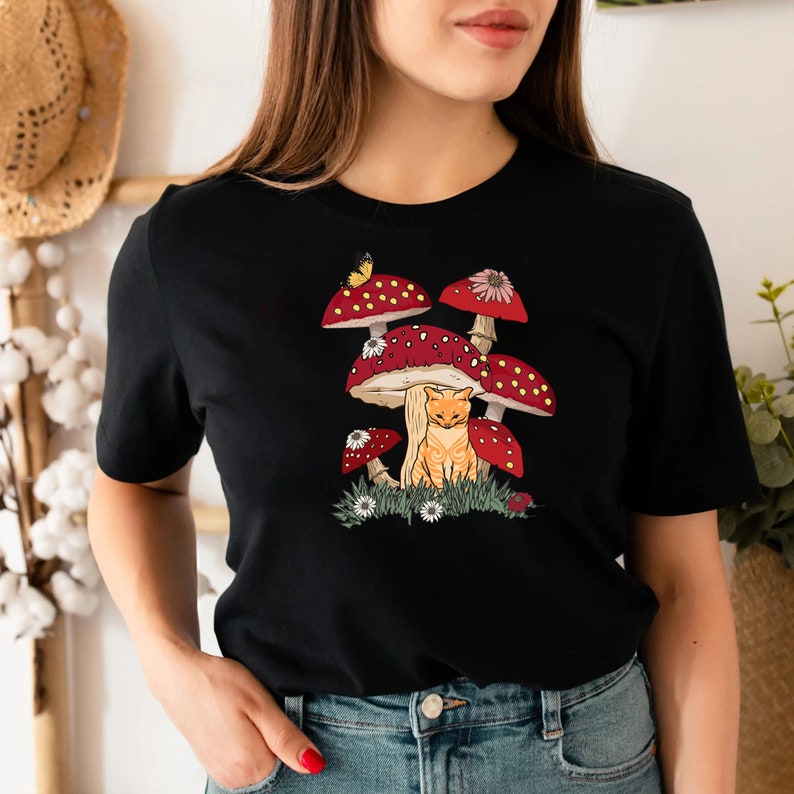 K&ouml;nnte beinhalten: Schwarzes T-Shirt mit einer skurrilen Illustration einer orangefarbenen Katze unter roten Pilzen mit wei&szlig;en Punkten. Ein Schmetterling und Blumen sind ebenfalls im Design enthalten. Das Shirt besteht aus einem weichen Material.