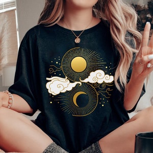 May include: A black t-shirt featuring a gold sun and moon design. The design incorporates clouds and stars in a celestial theme.