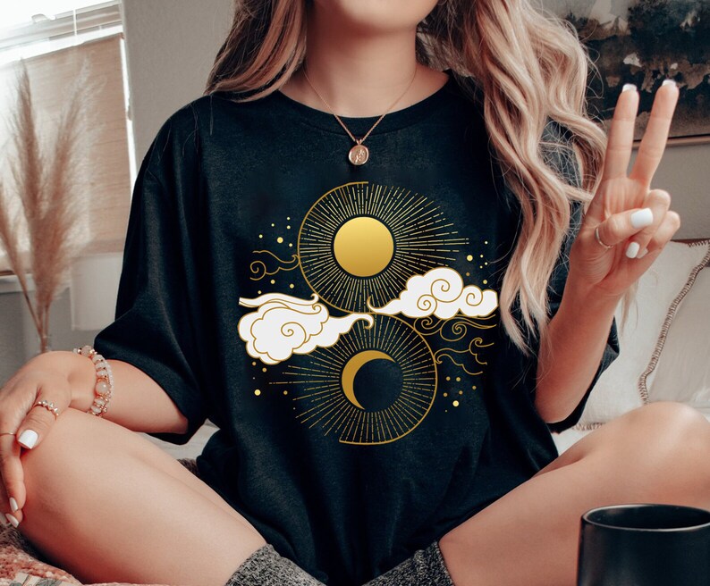 May include: A black t-shirt featuring a gold sun and moon design. The design incorporates clouds and stars in a celestial theme.