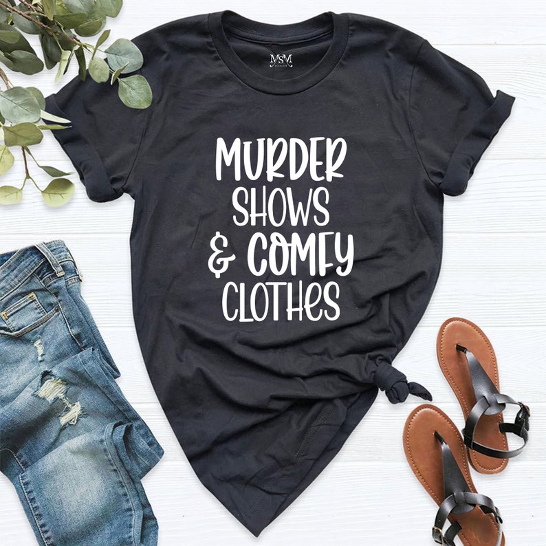 May include: Black t-shirt with the text "MURDER SHOWS & COMFY CLOTHES" in white. The shirt is styled with a knot at the bottom. A pair of brown sandals and a pair of blue jeans are also in the image.