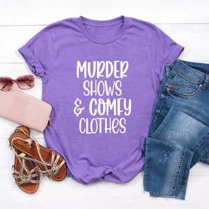May include: A purple t-shirt with the text "MURDER SHOWS & COMFY CLOTHES" in white, paired with blue jeans, a pink wallet, brown sandals, and pink sunglasses. The items are arranged on a white wooden surface.