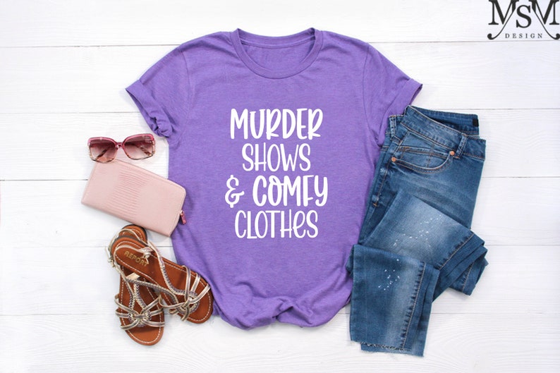 May include: A purple t-shirt with the text "MURDER SHOWS & COMFY CLOTHES" in white, paired with blue jeans, a pink wallet, brown sandals, and pink sunglasses. The items are arranged on a white wooden surface.