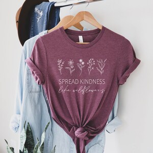 Op de afbeelding: Een maroon t-shirt met een witte afbeelding van wilde bloemen en de tekst "SPREAD KINDNESS like wildflowers".