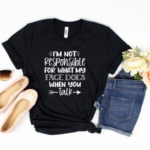 以下が含まれることがあります： I'M NOT RESPONSIBLE FOR WHAT MY FACE DOES WHEN YOU TALKという白い文字がプリントされた黒い半袖Tシャツ。ピンクのバレエシューズ、ダークブルーのデニムショートパンツ、茶色のサングラス、そして桃色の花の小さなブーケが添えられています。