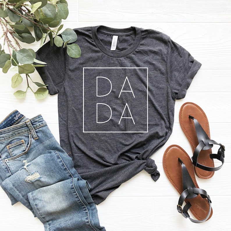May include: A dark gray t-shirt with a white square graphic that says "DA DA" on it. The shirt is laid out on a white surface with a pair of brown sandals and a pair of blue jeans.