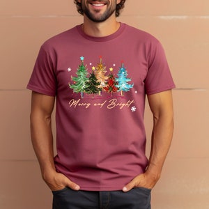 Pu&ograve; includere: Una maglietta color granata con un disegno di albero di Natale e il testo "Merry and Bright".