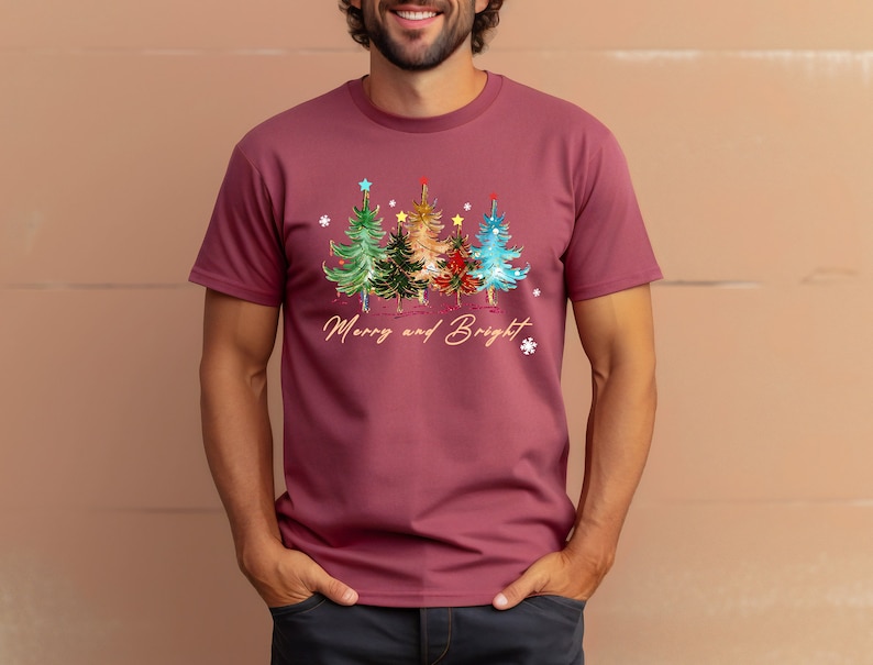Pu&ograve; includere: Una maglietta color granata con un disegno di albero di Natale e il testo "Merry and Bright".