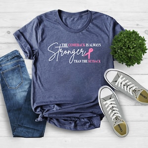 May include: A navy blue t-shirt with the text "The Comeback Is Always Stronger Than The Setback" in white and pink. The shirt is paired with blue jeans and gray sneakers.
